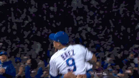 Baseball player celebrating a victory on the field as teammates rush in, with cheering fans in the background.
