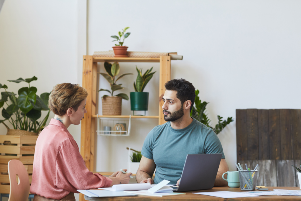 Two people talk across desk with laptop and papers.