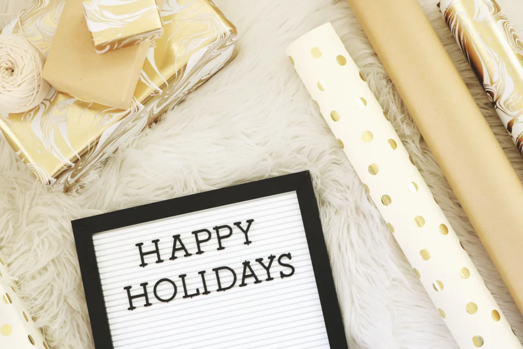 A letter board with the words “Happy Holidays” surrounded by gold gift wrap, ribbon, and presents.