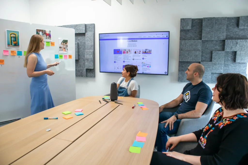 A woman presents ideas on a whiteboard to a team during a meeting with sticky notes and a screen display.