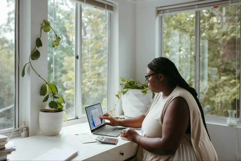 Woman working on a laptop by a large window with natural light.