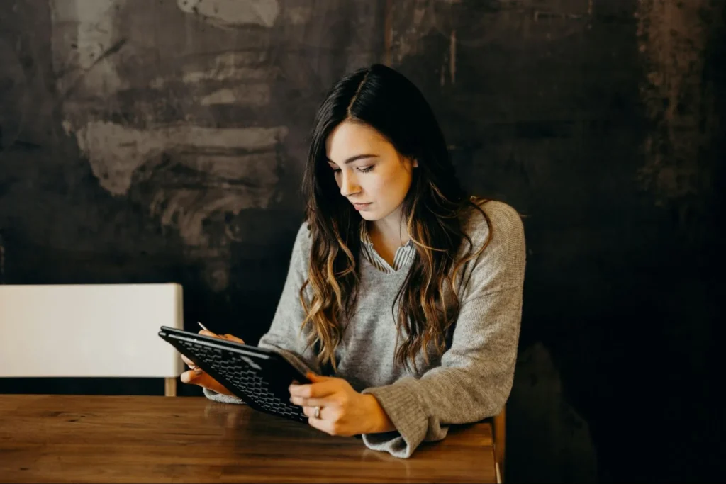 A woman sits at a wooden table reading or working on a tablet.