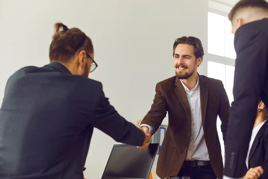 A businessman smiling and shaking hands with a new team member in an office, representing a successful onboarding or partnership.
