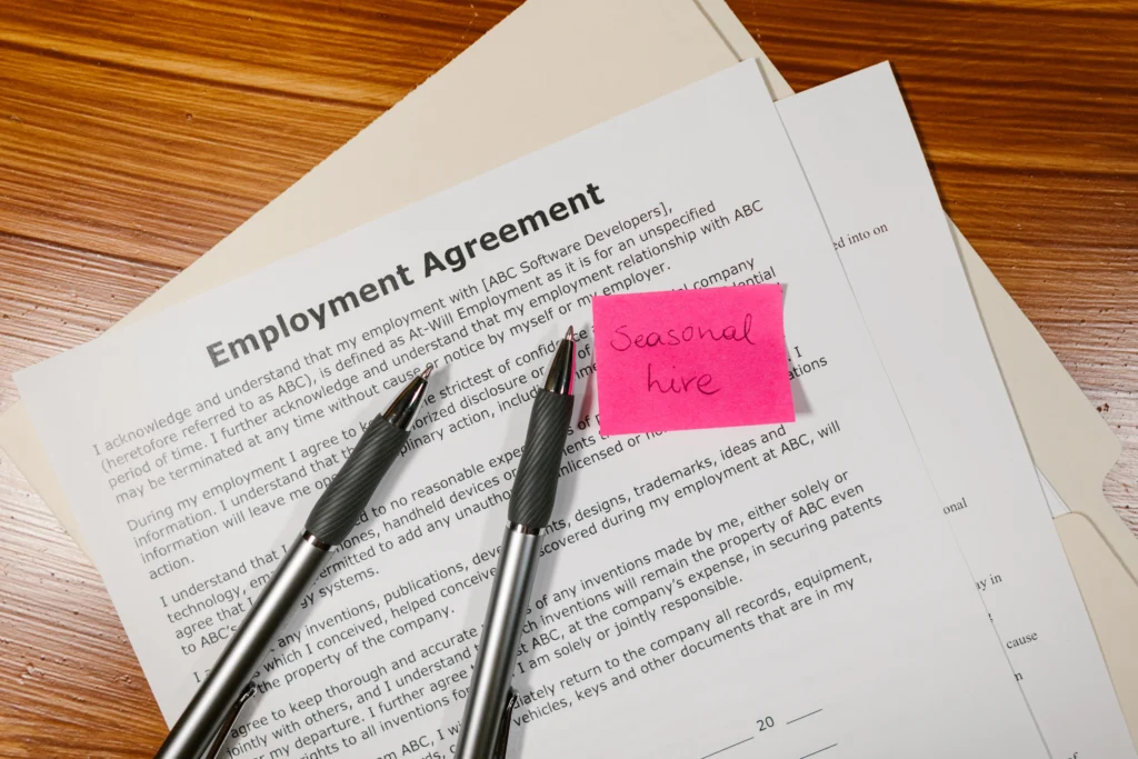 Employment agreement with pink sticky note reading “seasonal hire” beside two pens.