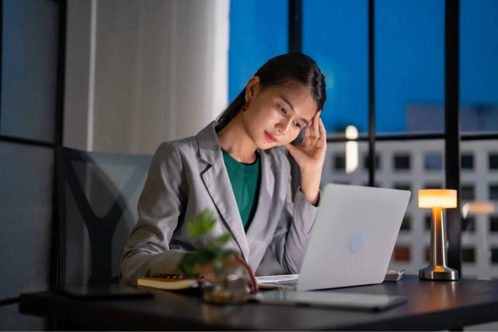 A woman in a blazer works on her laptop at night, appearing focused under warm desk lighting.