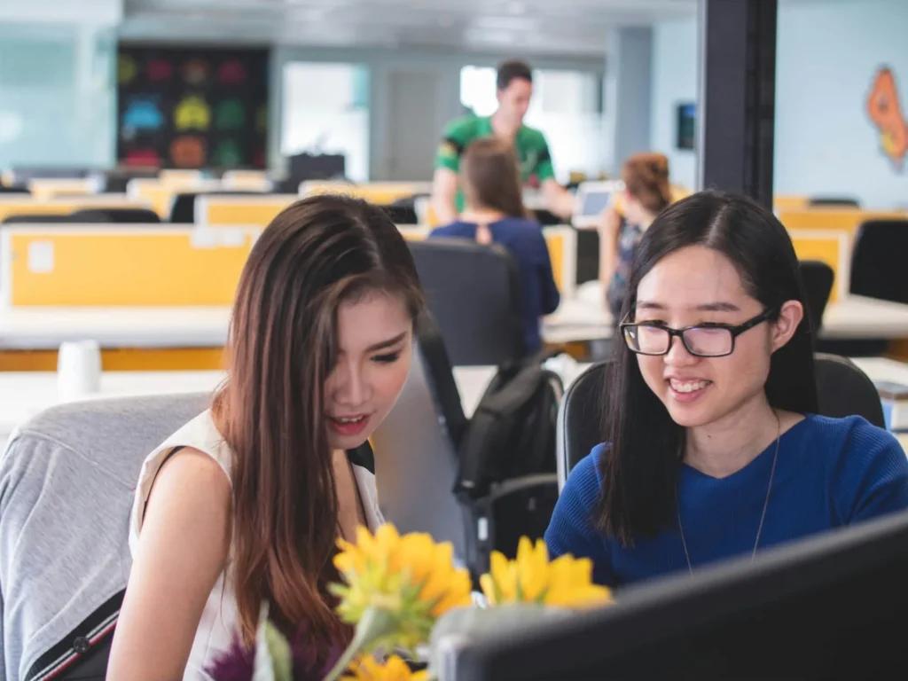 Two women collaborating and smiling while working on a computer in a modern office.
