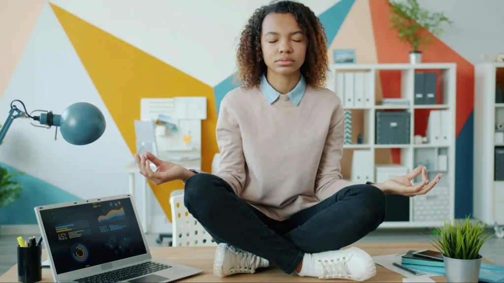 Person sitting cross-legged on a desk in an office, meditating with a laptop and plants nearby.