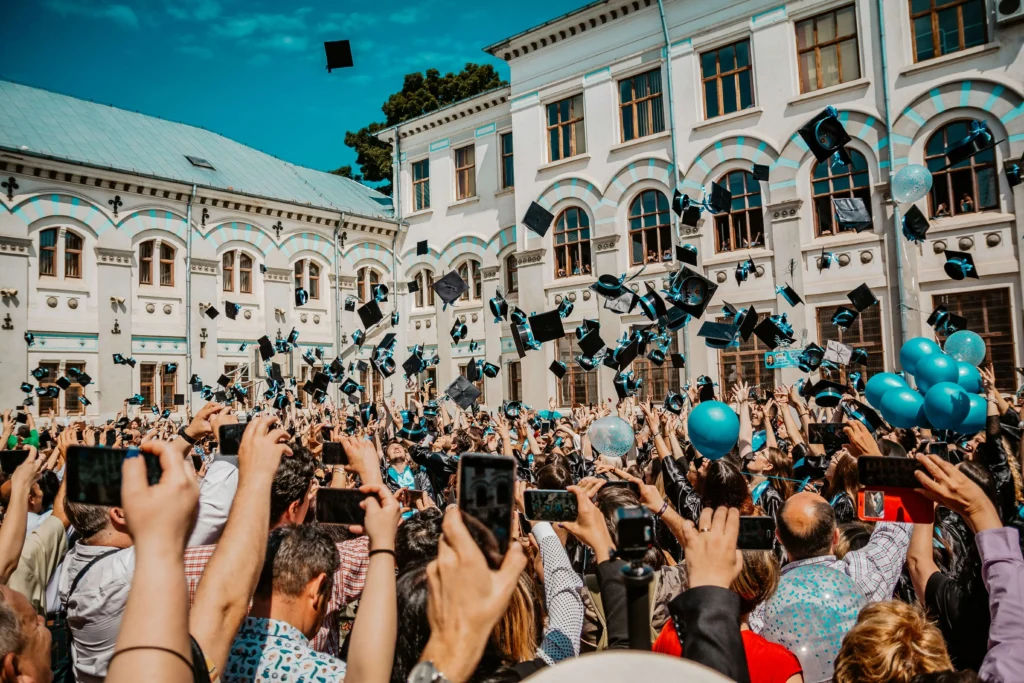Crowd celebrating a graduation ceremony with graduates tossing caps into the air outside a university building.