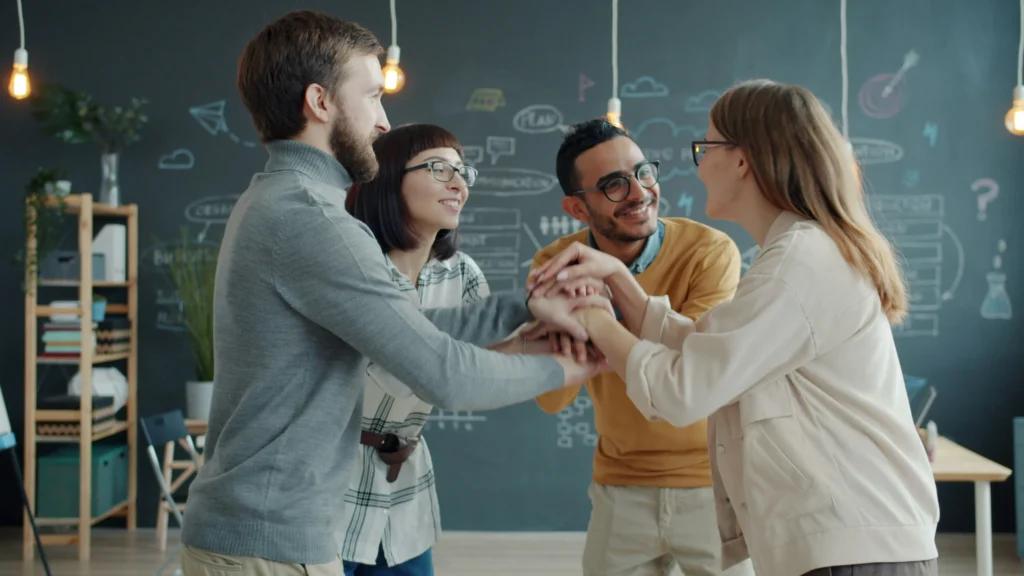 A diverse group of four people stand in a circle with hands joined, smiling and laughing. They're in a modern office with a chalkboard wall and hanging lights, conveying teamwork and positivity.