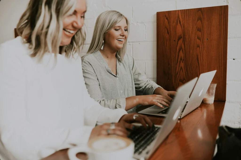 Two women are seated at a table, working on laptops while discussing ideas.
