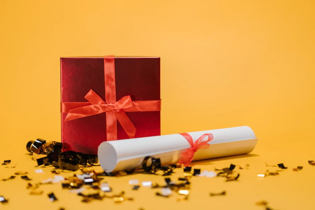 Red gift box and rolled certificate with confetti on a yellow background, representing employee recognition and rewards.