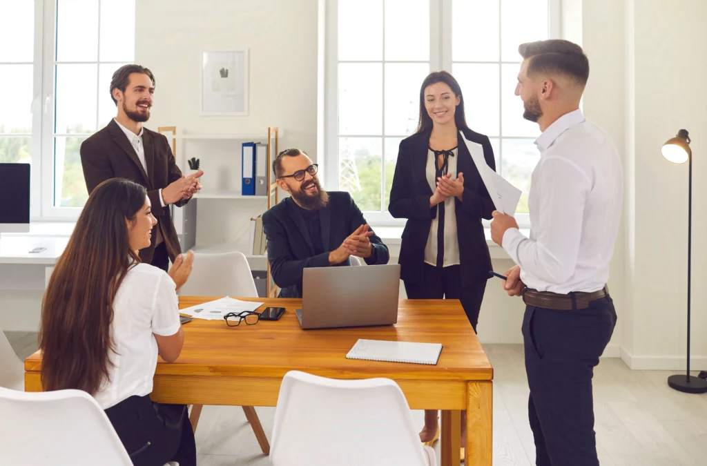 Team of coworkers applauding a colleague giving a presentation in a modern office, showing employee recognition in action.