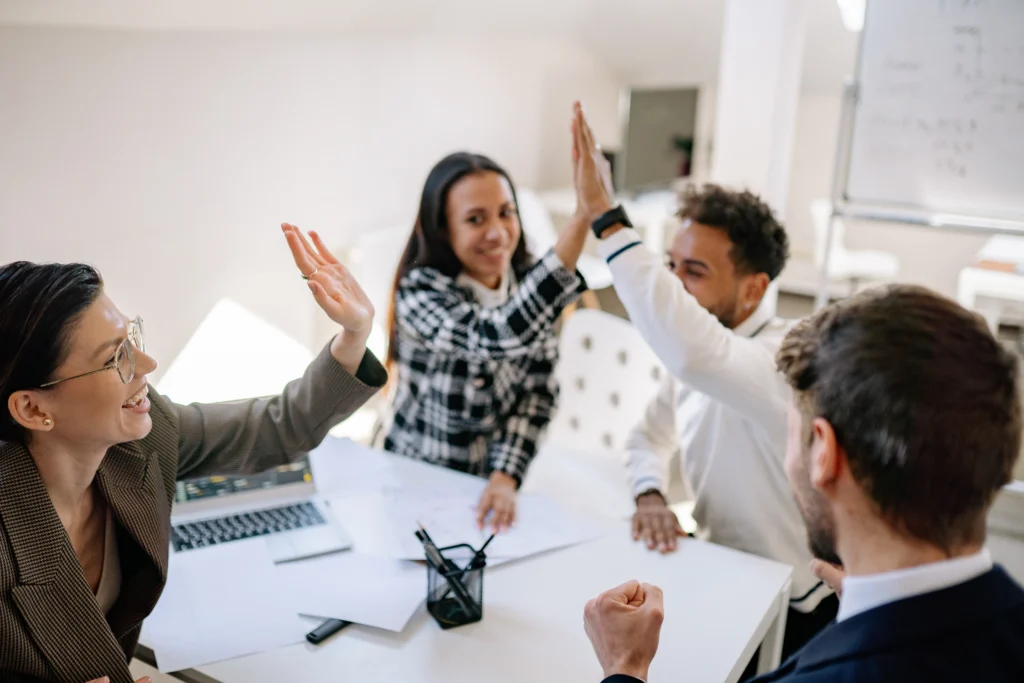 Group of smiling employees high-fiving in celebration during a meeting, symbolizing teamwork and employee recognition.