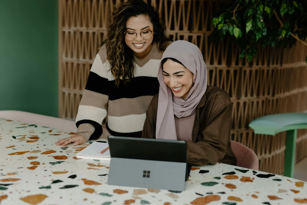 Two women wearing hijabs are seated at a table, working together on a laptop, sharing ideas and insights.