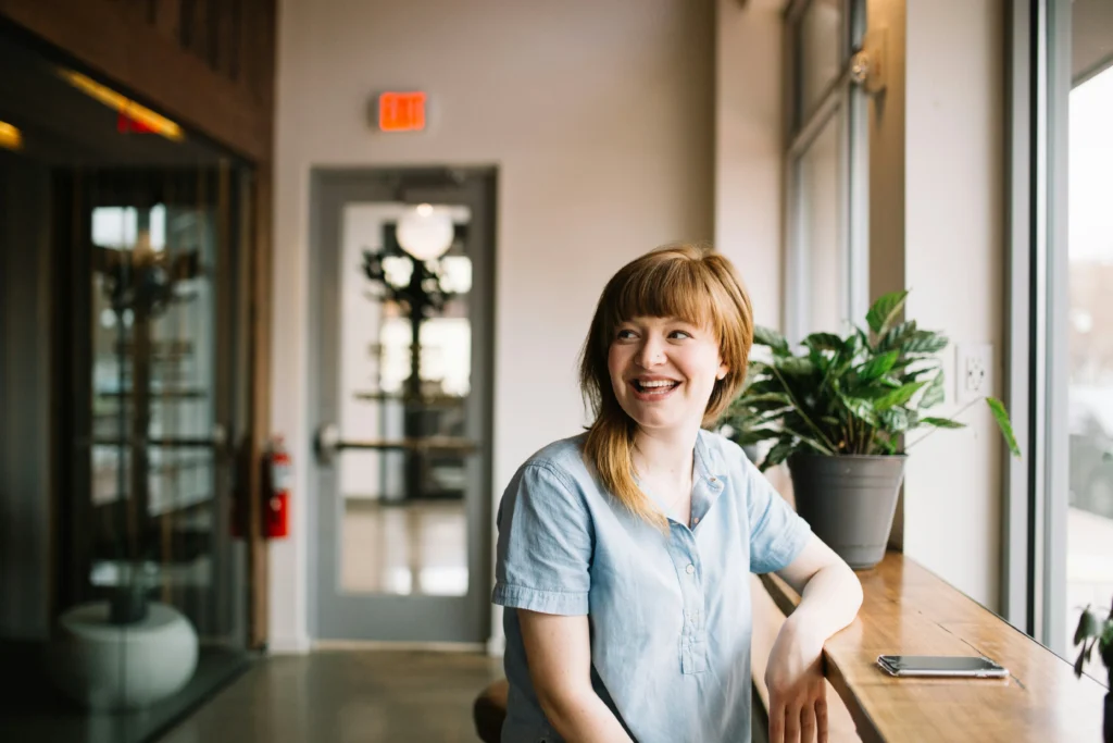 A smiling woman sitting by a window in a modern workspace, representing happy remote workers.