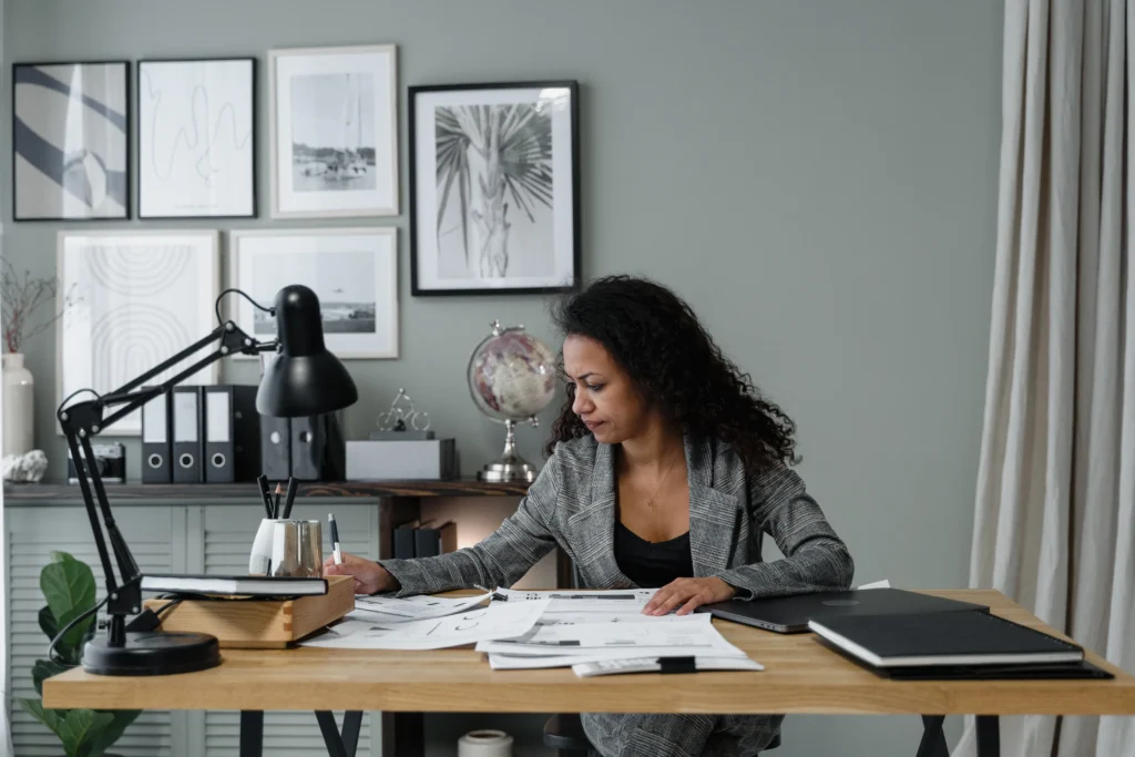 A woman in a gray blazer works at a wooden desk covered with papers, a lamp, and notebooks. Behind her, a wall displays framed art. The scene is calm and focused.