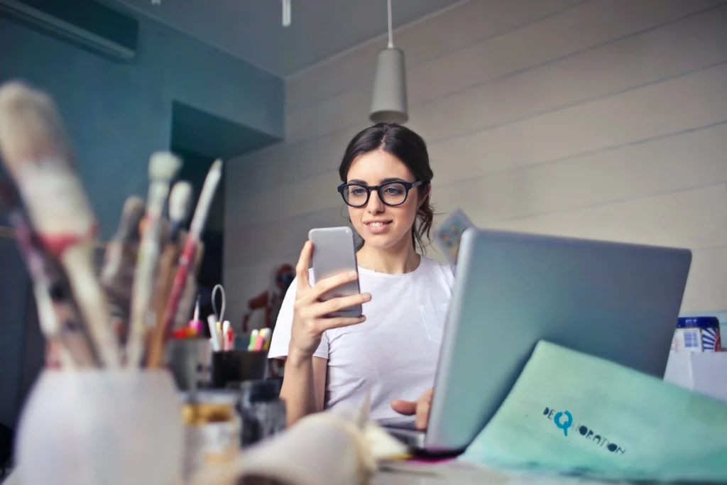 A woman wearing glasses sits at a desk, focused on her phone while engaged in her work.