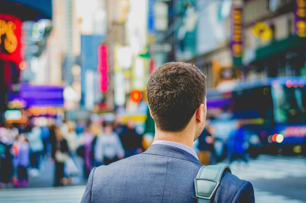A businessman in a suit, seen from behind, walking in a busy metropolitan area with tall buildings and blurred lights.