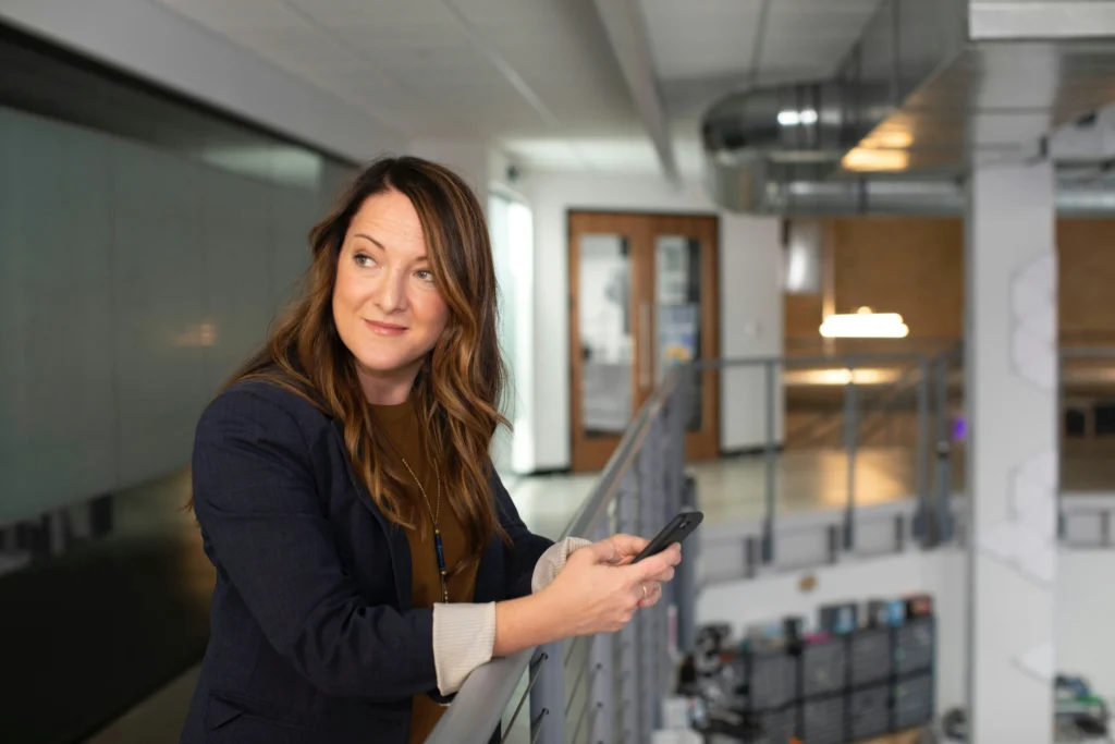 A professional woman standing on a mezzanine level in a modern office building, holding a mobile phone and looking into the distance.