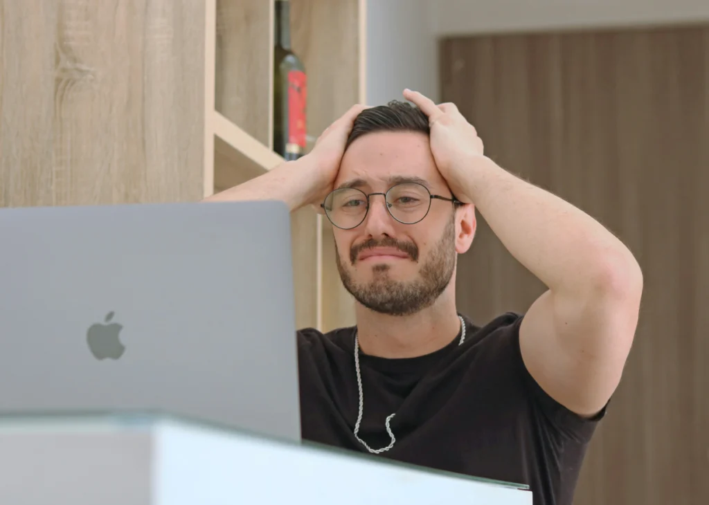 A man with glasses looking stressed or frustrated while sitting in front of a laptop, holding his head with both hands.