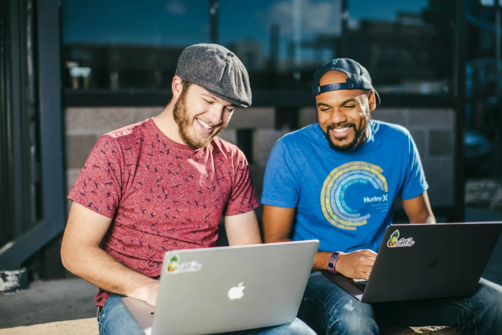 Two men sitting on a bench, each using a laptop, engaged in conversation or work.
