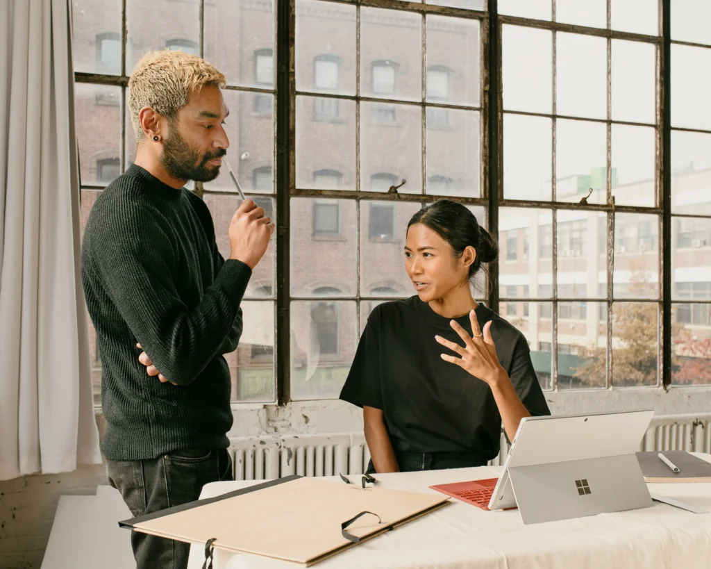 A man and woman engaged in conversation at a table, with a laptop open between them.