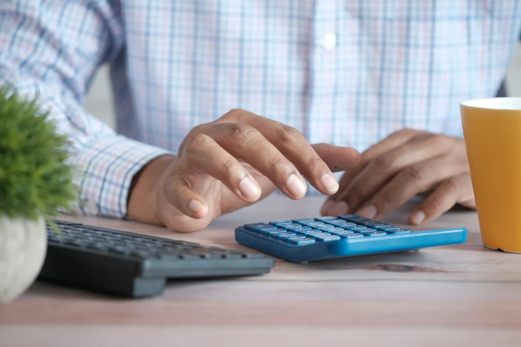 A close-up of a person's hands using a blue calculator on a wooden desk next to a keyboard and a coffee mug, for finance or accounting.