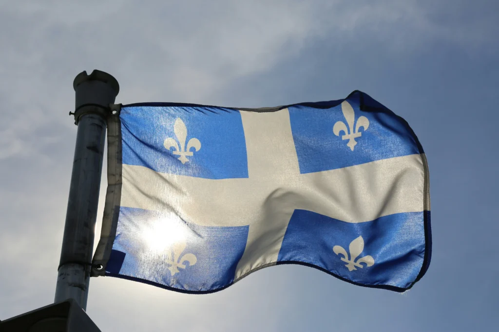 The white and blue flag of Quebec, featuring four fleurs-de-lis, blowing in the wind against a bright sky.