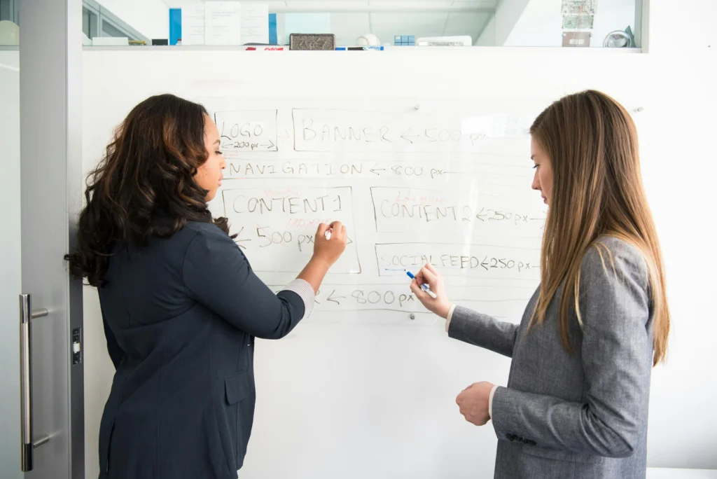 Two businesswomen writing and sketching design specifications, content layout, and navigation notes on a large glass whiteboard.