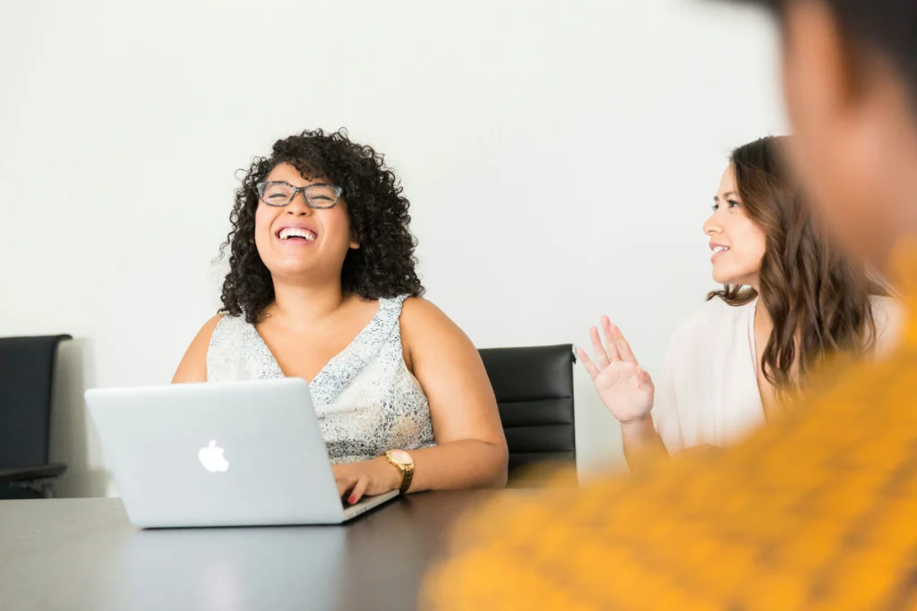 A woman in glasses laughing joyfully while seated at a dark table with a laptop during an informal office meeting.