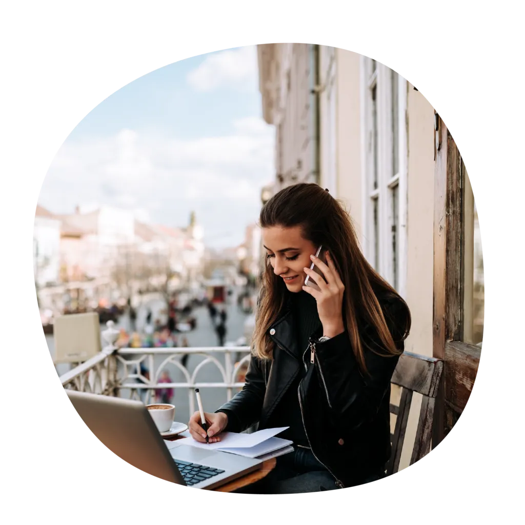 Smiling woman working on a balcony with a laptop, writing notes, and talking on the phone.