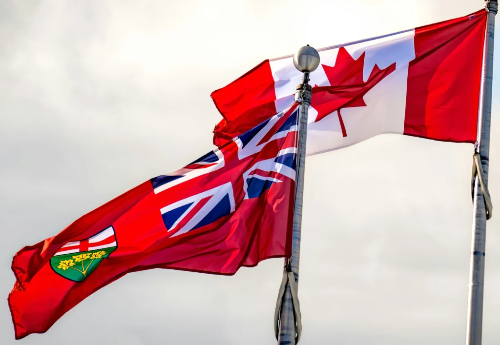 Canadian flag and Ontario provincial flag flying side by side on flagpoles against a cloudy sky.