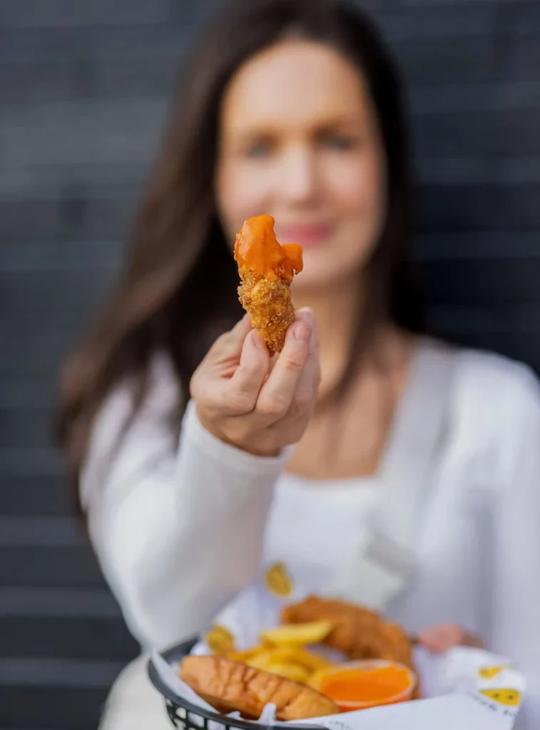 A woman in white holds out a fried chicken tender with sauce. In her other hand, she holds a basket with fries and an orange slice. The background is a dark blurred wall.