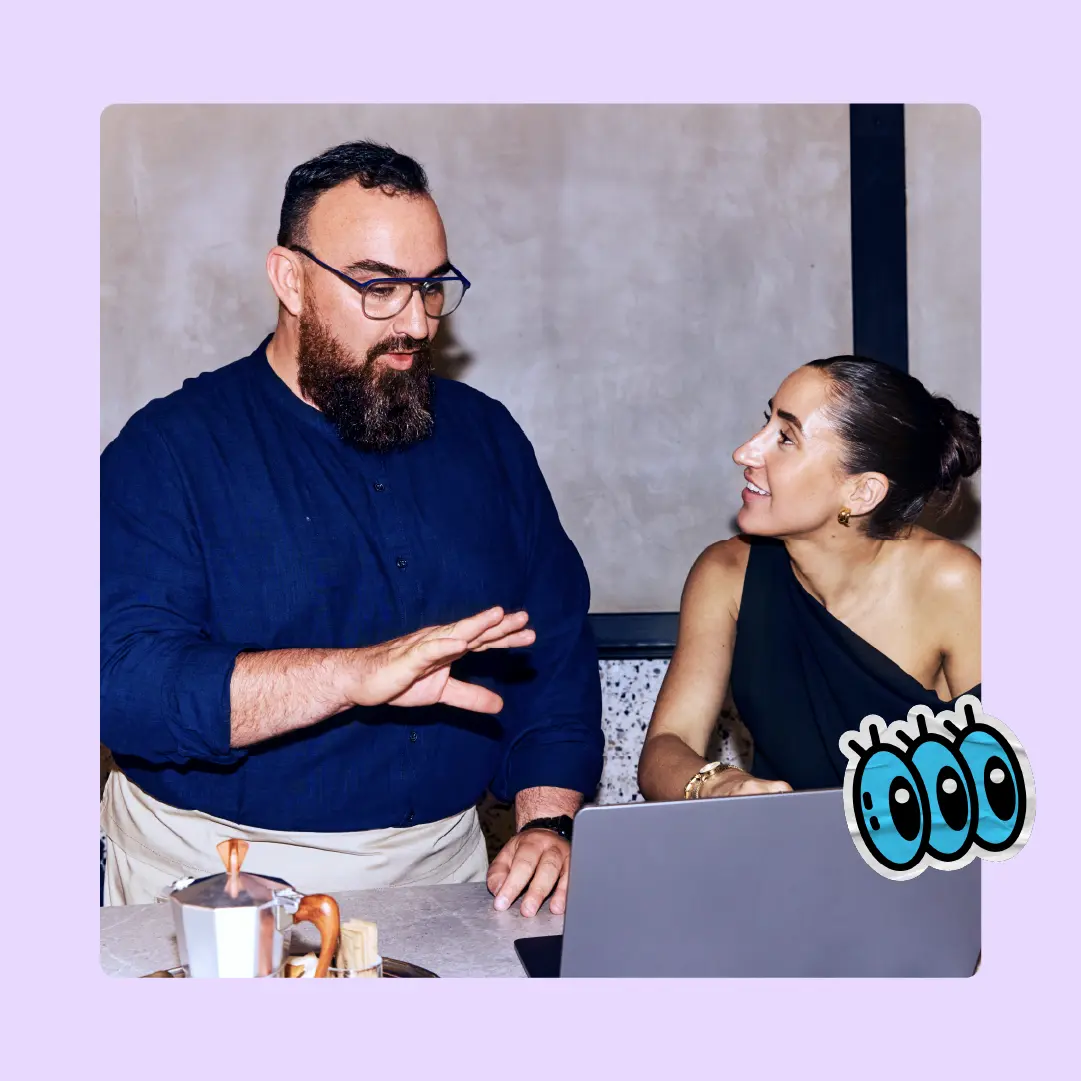 A bearded man in a navy shirt gestures while speaking to a smiling woman in a black dress. They sit at a table with a laptop, creating a collaborative and engaged atmosphere.