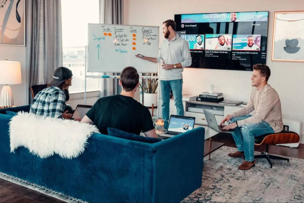 Four men in a modern office setting have a casual meeting. One stands by a whiteboard with tasks listed, while others sit with laptops, looking engaged.