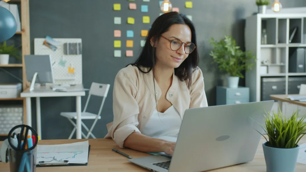 A woman in glasses works on a laptop at a desk, surrounded by plants, charts, and colorful sticky notes. The space feels modern and organized.