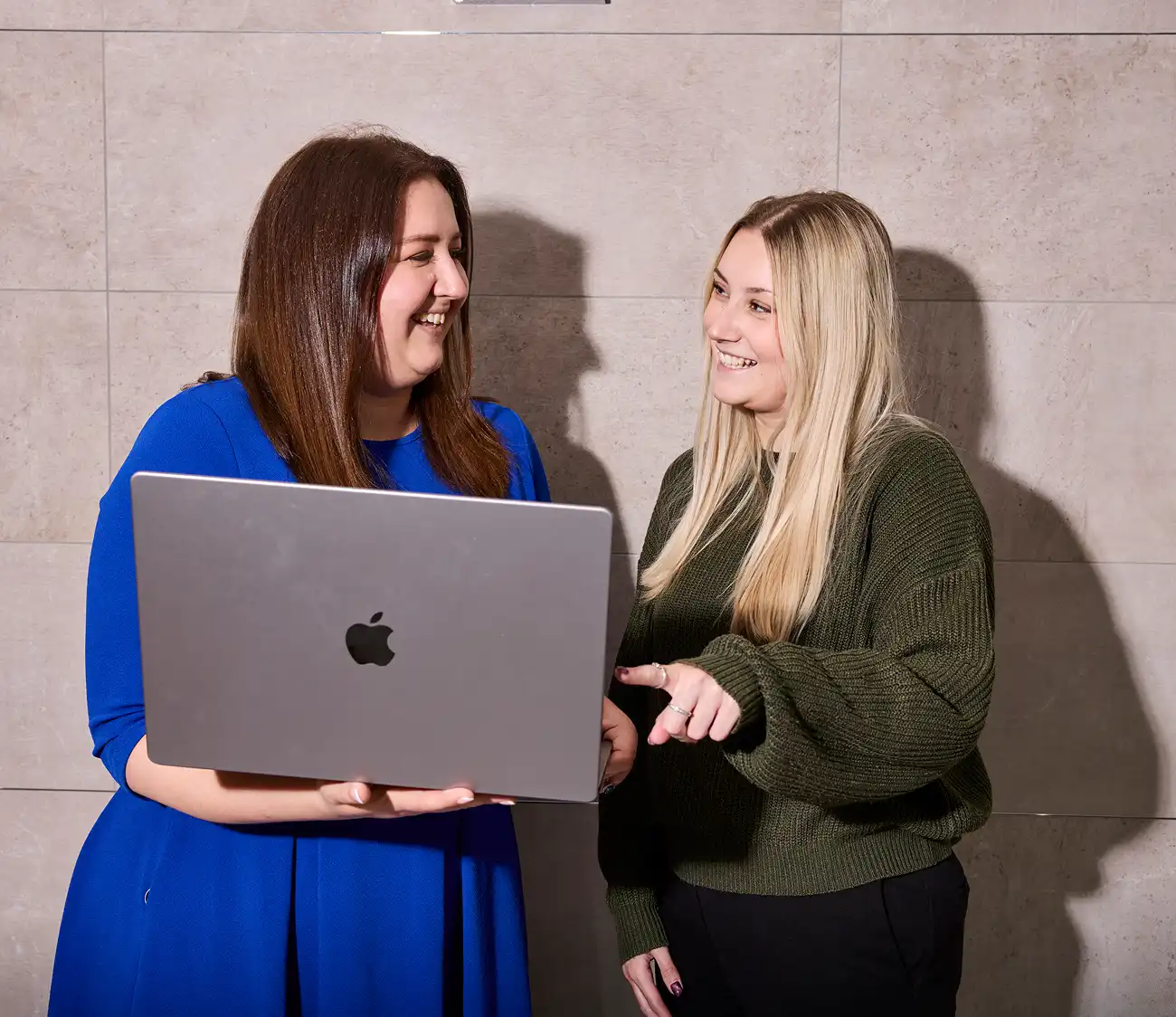 Two women smiling and looking at a laptop together. One wears a blue dress, the other a green sweater. They stand against a light-colored tiled wall.