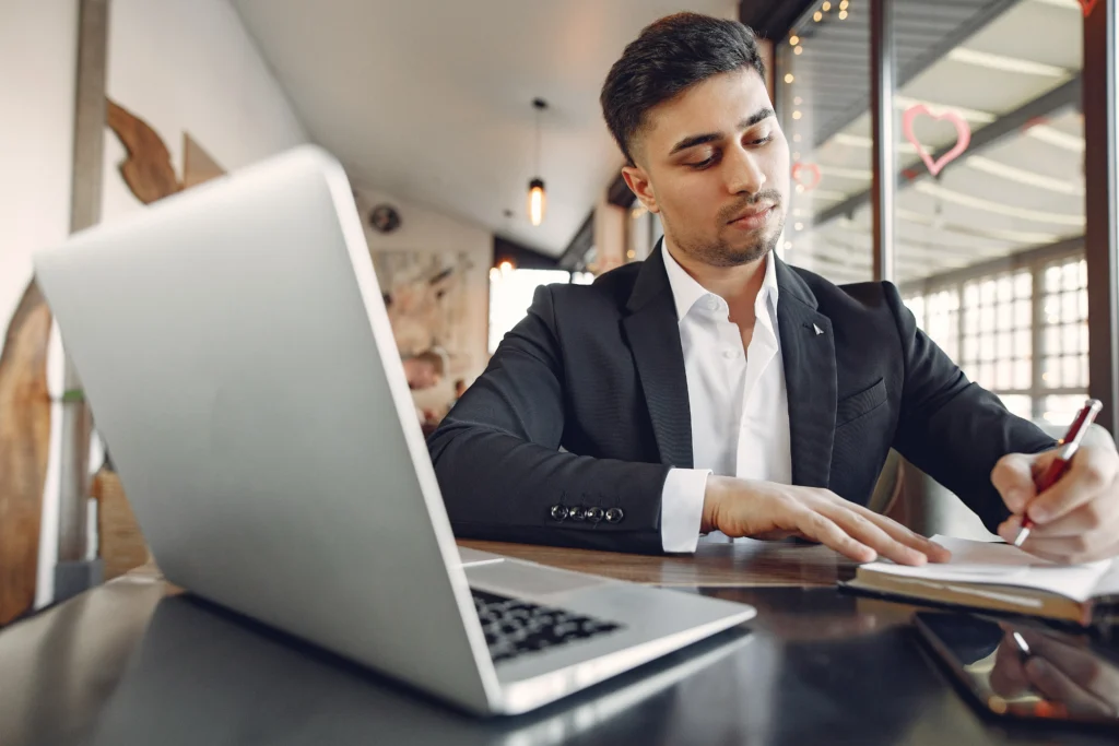 A businessman in a suit writing in a notebook while working on a laptop.