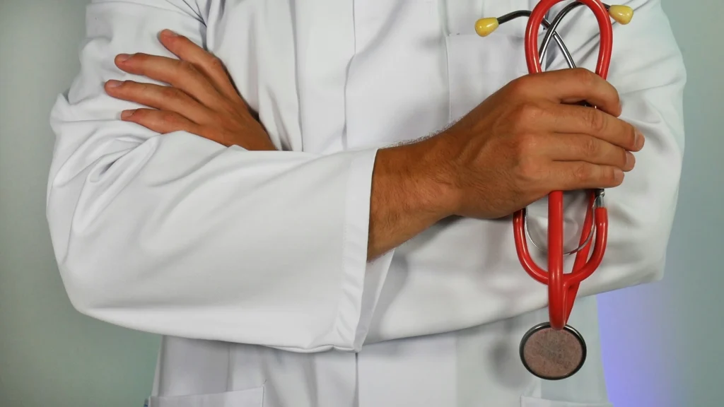 A doctor in a white coat holding a red stethoscope, arms folded confidently across the chest. The image conveys professionalism and care in healthcare.