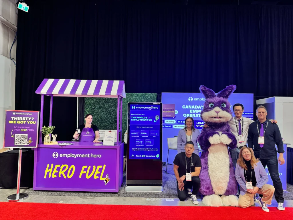 A group of smiling people poses with a large purple mascot, showcasing a fun and lively atmosphere at an event.