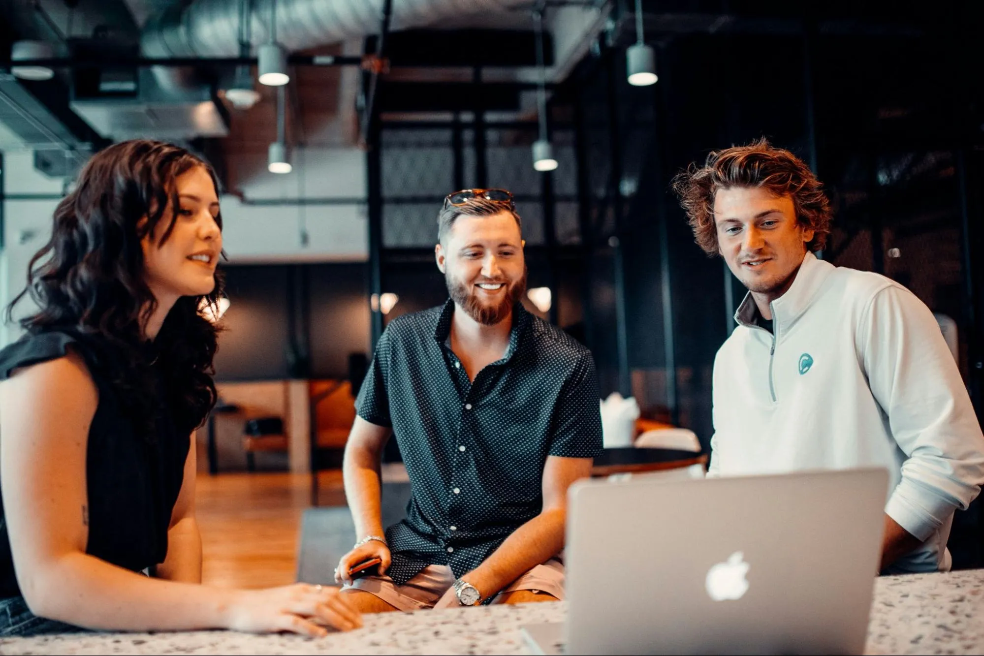 Three smiling colleagues gathered around a laptop in a modern office, representing a team collaborating and learning how to integrate AI at work.