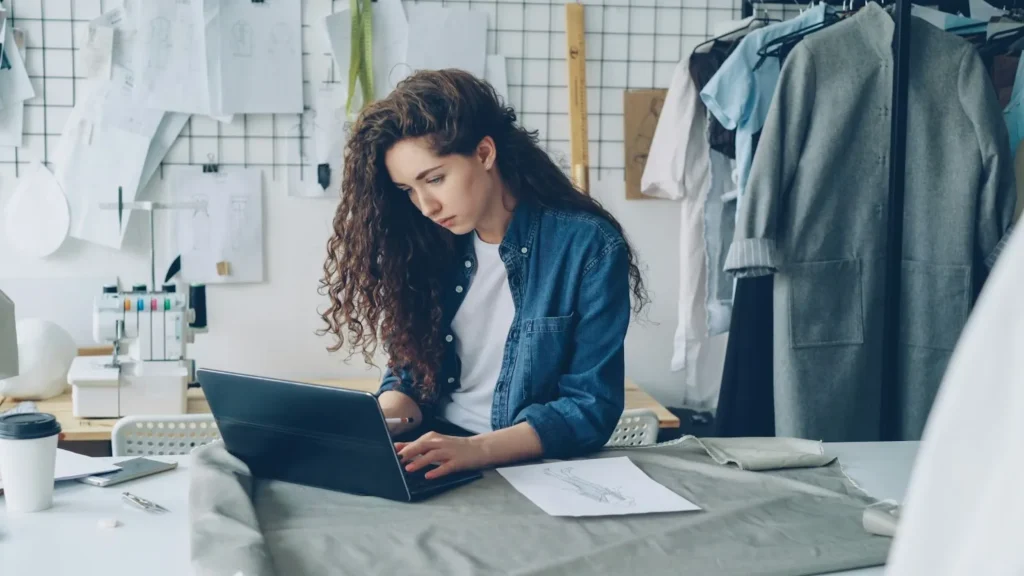 A female fashion designer with curly hair using a laptop at a workspace surrounded by fabric sketches, a sewing machine, and clothing racks in a bright studio.