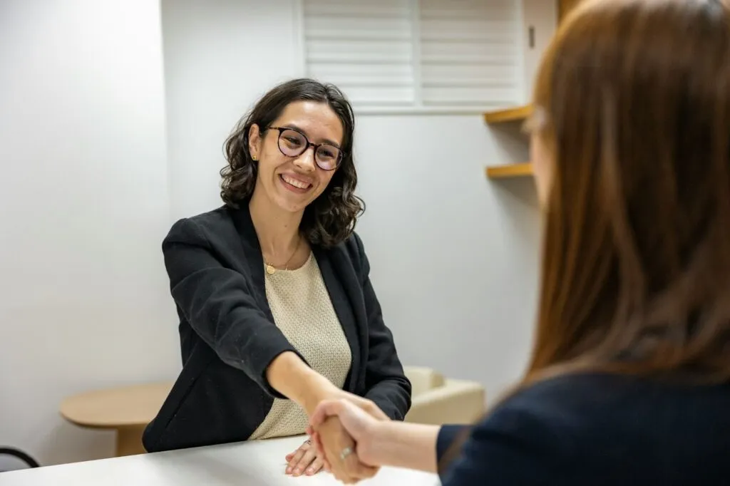 A smiling hiring manager shaking hands with a candidate across a desk, highlighting the interview stage essential to any successful hiring strategy.