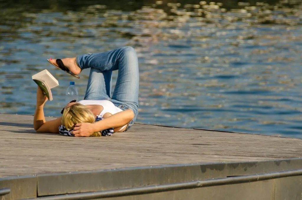 A woman relaxing and reading a book on a wooden dock by the water, highlighting personal sabbaticals or extended mental health breaks as potential scenarios for taking leave without pay.
