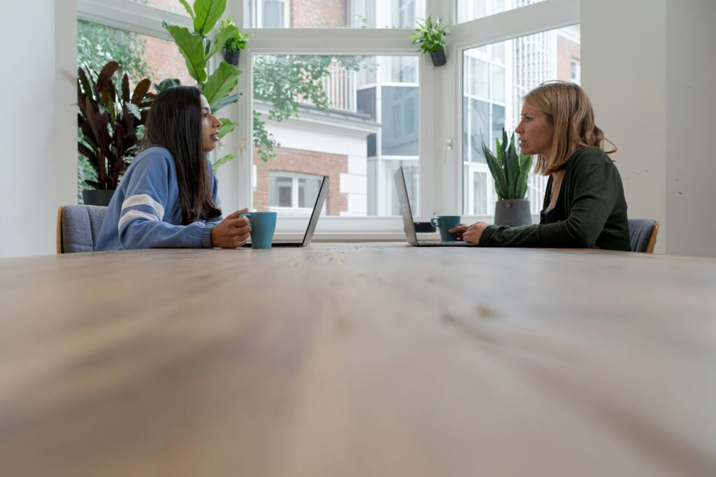 Two women engaging in a serious one-on-one meeting in a plant-filled office. They are seated opposite each other at a large wooden table with laptops and mugs, depicting a manager and employee preparing to discuss performance feedback and coaching, aligned with one-on-one meeting questions for managers.