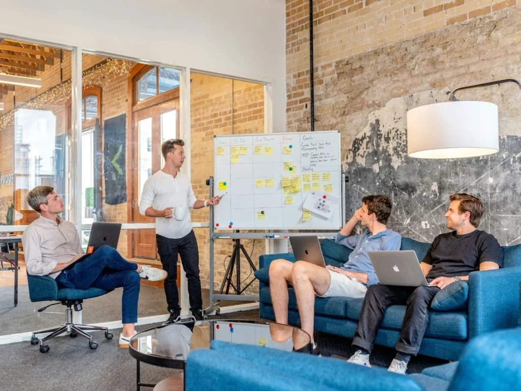 A team member presenting at a whiteboard with sticky notes while colleagues listen from a sofa, highlighting clear visual presentations as a way to resolve communication barriers in the workplace.
