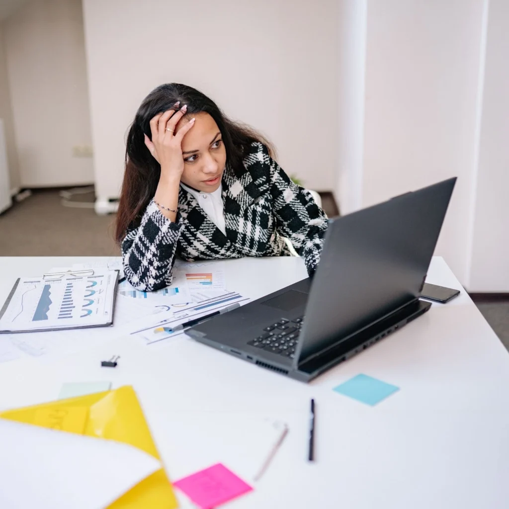 Woman in a checkered jacket looks stressed, leaning on a white desk cluttered with colorful papers, a laptop, and charts, conveying frustration.