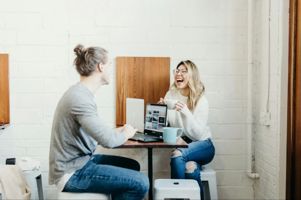 A woman laughs joyfully while holding a mug and chatting with a colleague at a small cafe table equipped with laptops and coffee.