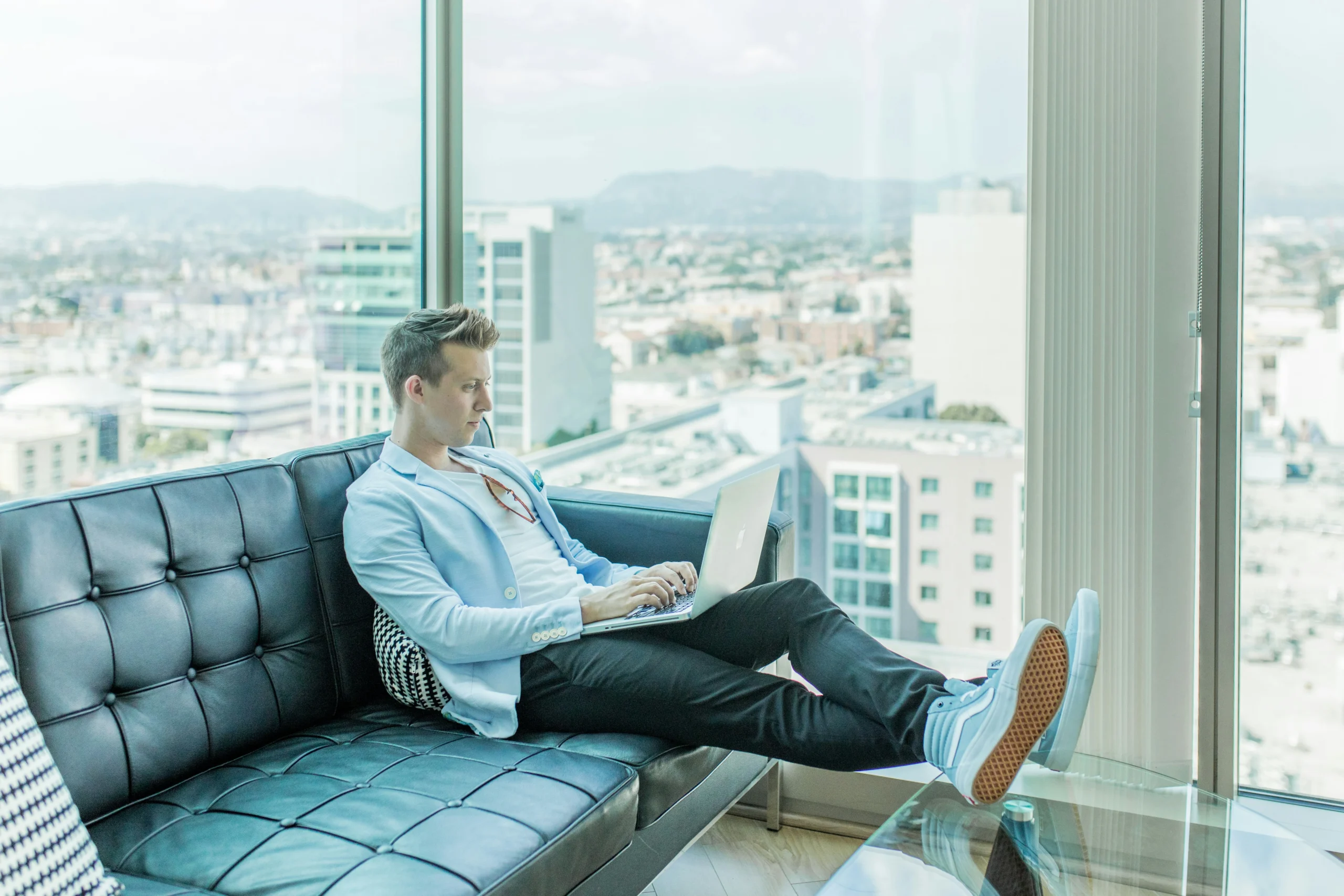 A man in a light blue blazer works on his laptop while lounging on a black leather sofa in a high-rise office with a sprawling city view.