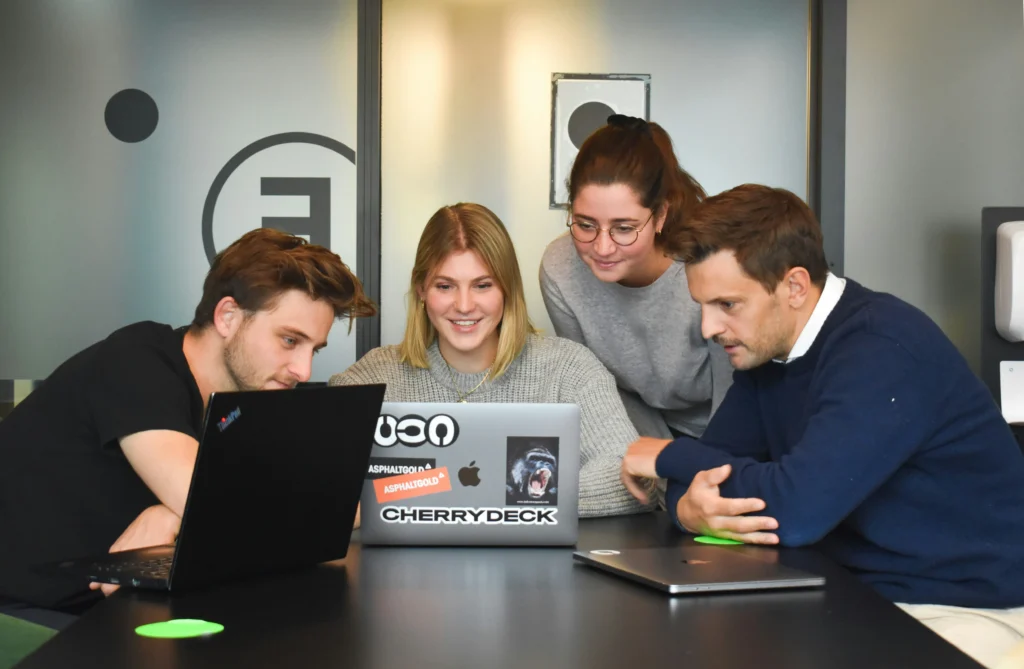 Four people gathered around laptops in an office, collaborating and smiling. The atmosphere is focused yet relaxed, suggesting teamwork.